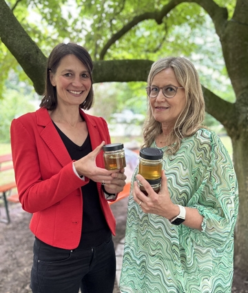 Ministerin Miriam Staudte mit Imkerin Wiebke Langefeld stehen im Garten des Ministeriums, sie halten Honiggläser in der Hand.