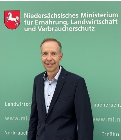 Dr. Jörg Baumgarte vor einer grüner Wand mit Ministeriumsaufschrift.
